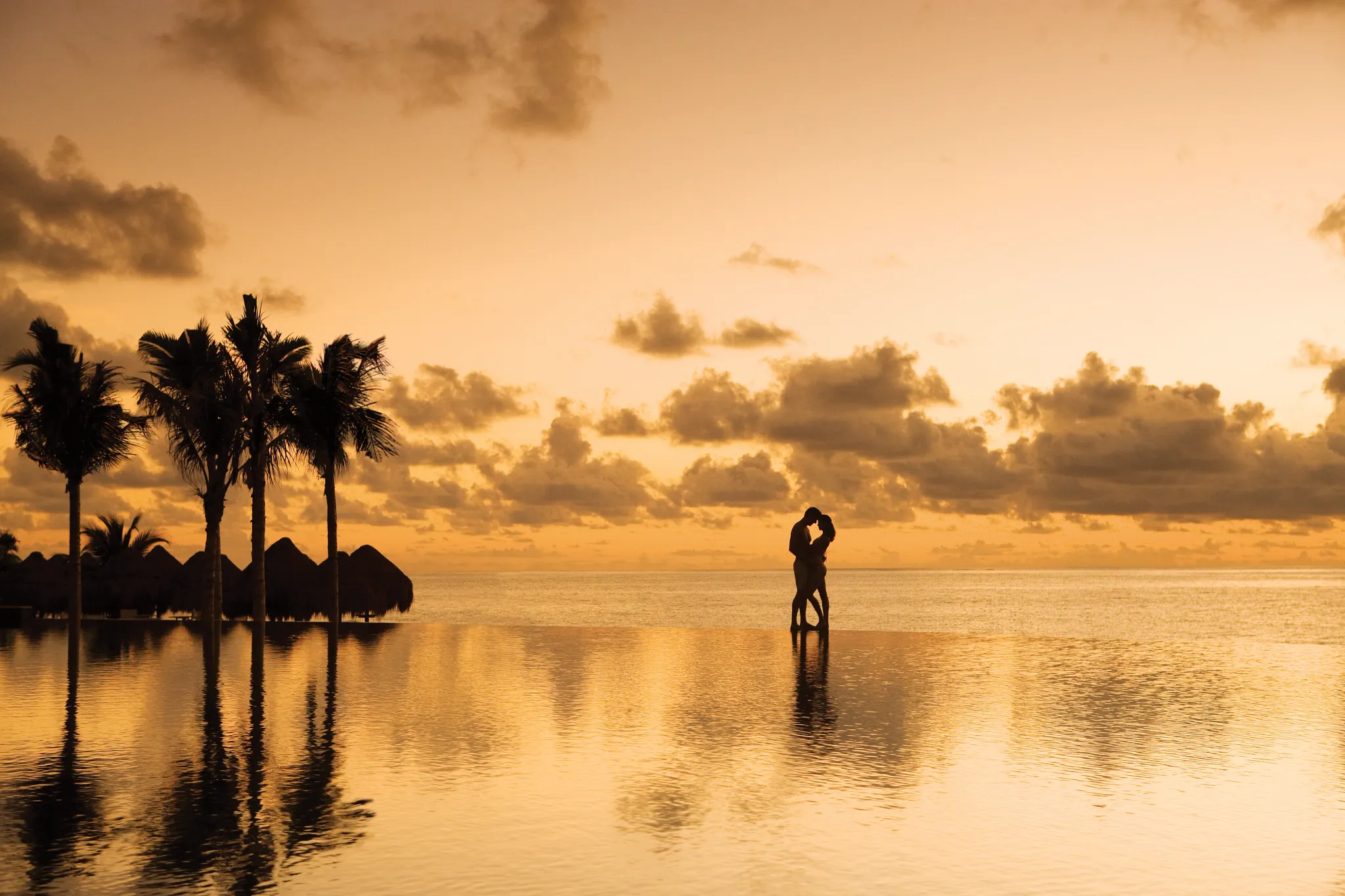 Couple embracing at sunset by an infinity pool at Dreams Riviera Cancun Resort & Spa with ocean views.