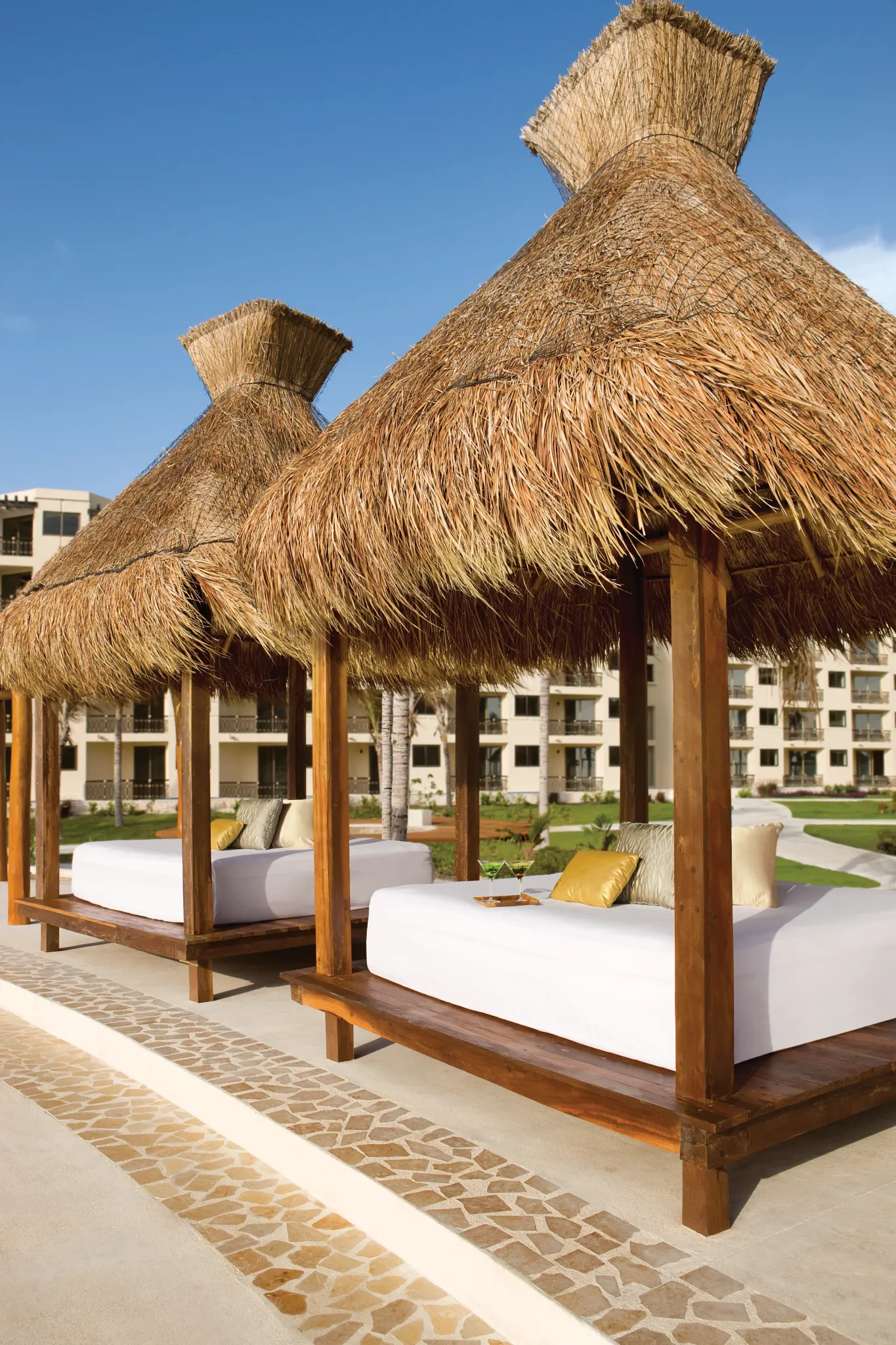 Thatched-roof daybeds with cushioned loungers at Dreams Riviera Cancun Resort & Spa outdoor relaxation area.